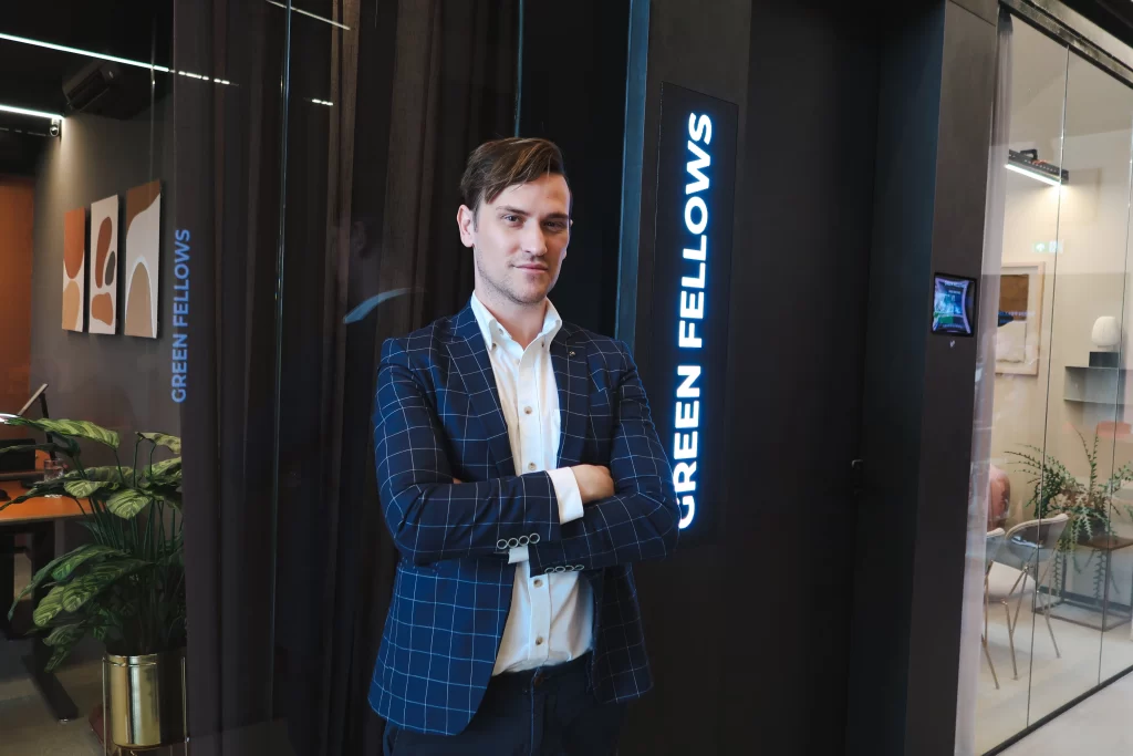 George in een blauwe blazer poseert met gekruiste armen voor de ingang van het Green Fellows-kantoor.