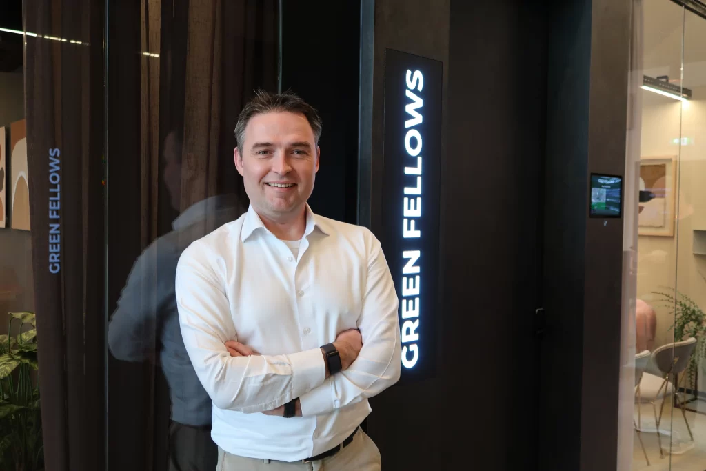 Arthur in een wit overhemd poseert voor het verlichte Green Fellows-logo bij de kantoorentree.