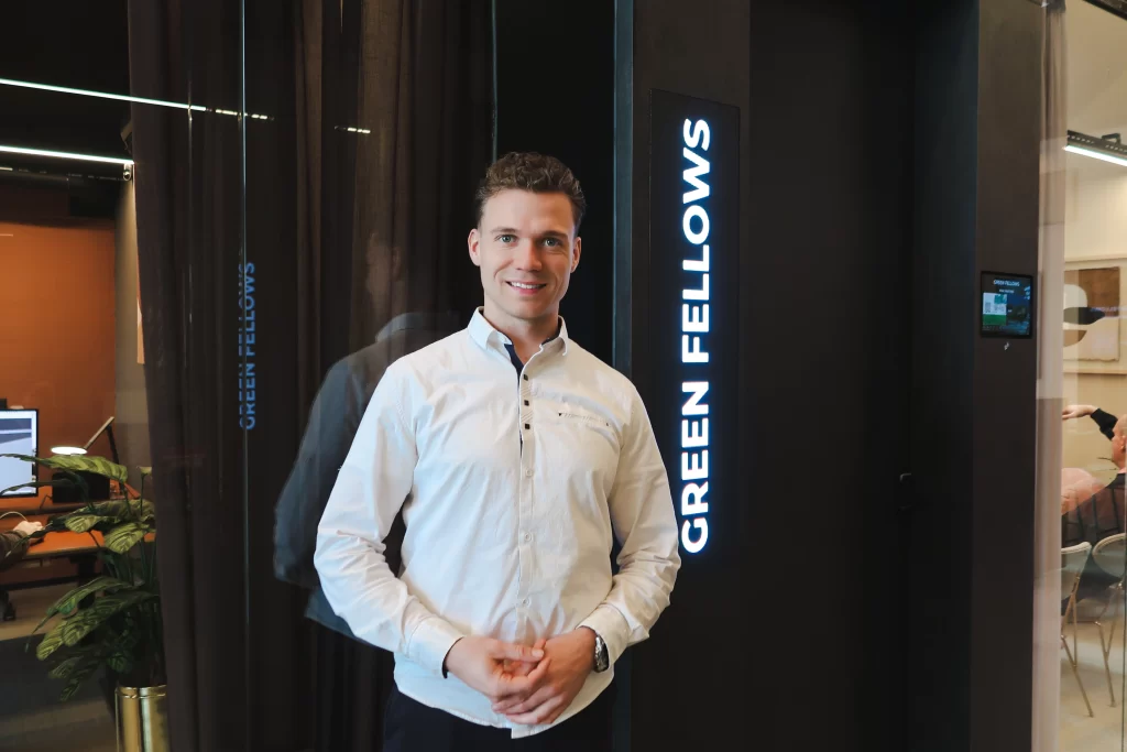 Matthijs in een wit overhemd poseert voor het verlichte Green Fellows-logo bij de ingang.