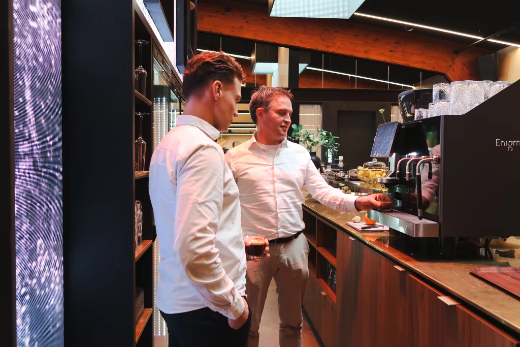 Matthijs en Arthur in een professionele keukenomgeving, in gesprek bij een houten bar met industriële uitstraling.