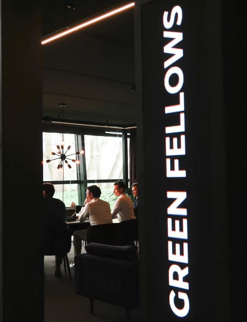 Sfeerbeeld van een overleg in een sfeervolle ruimte, met het verlichte Green Fellows-logo op de muur.