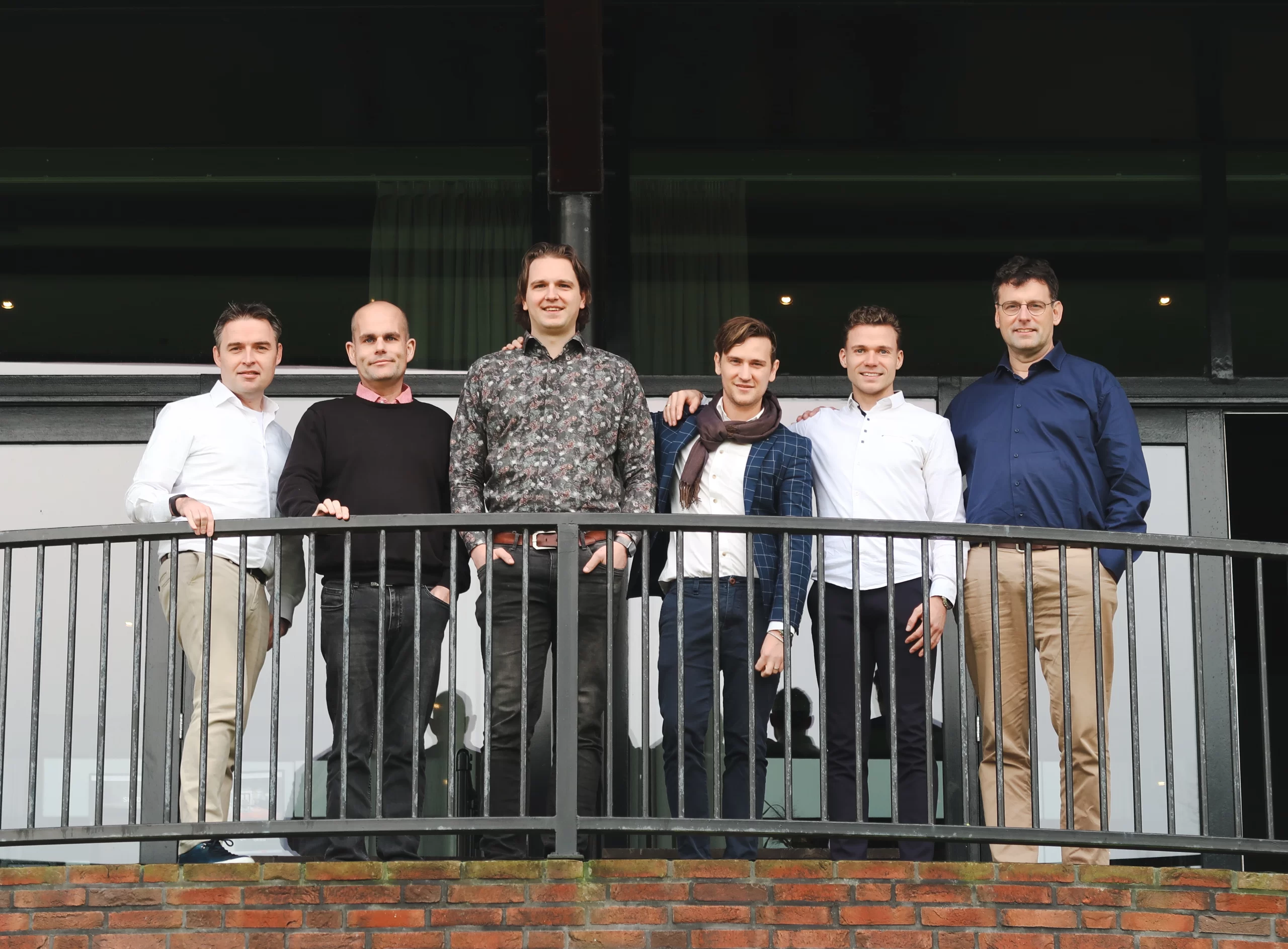 Het Green Fellows-team poseert buiten op een balkon, met een moderne kantoorgevel op de achtergrond.